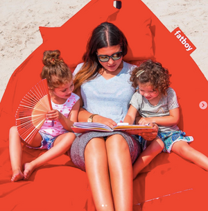 photo of a mother with her two children in a red hammock on the beach. The family is reading a book together.